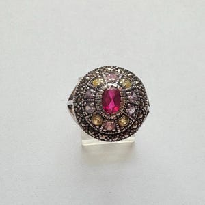May include: A silver-toned ring with a central oval ruby surrounded by smaller gemstones in pink, yellow, and purple hues. The ring has a detailed, textured band and a circular design around the central stone.