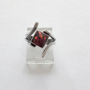 May include: A silver ring with a square, reddish-brown stone. The ring has a unique, abstract design with curved and angular elements. The stone has flecks of yellow and orange. The ring is displayed on a clear, rectangular stand.
