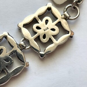 以下が含まれることがあります： 正方形のチャームが花柄のデザインで飾られたシルバー製のチャームブレスレットのクローズアップ。チャームには「925」の刻印があり、スターリングシルバー製であることを示しています。