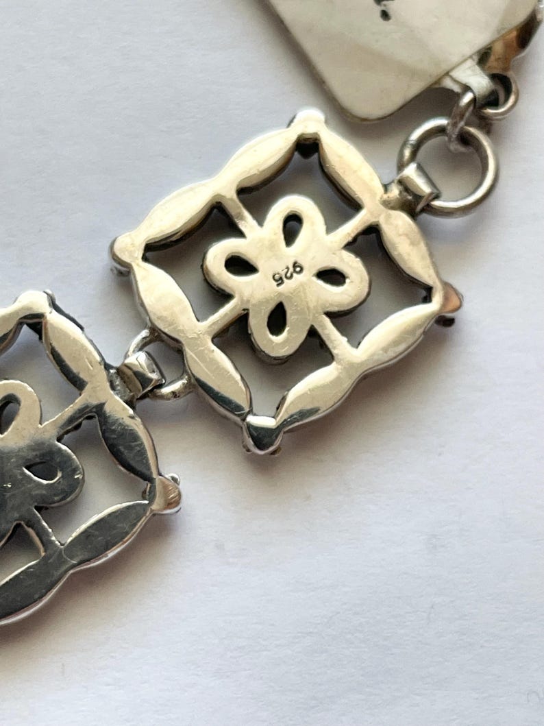 以下が含まれることがあります： 正方形のチャームが花柄のデザインで飾られたシルバー製のチャームブレスレットのクローズアップ。チャームには「925」の刻印があり、スターリングシルバー製であることを示しています。