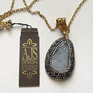 May include: A gold chain necklace with a pendant featuring a light blue agate stone surrounded by black crystals. The pendant is attached to a gold chain with a lobster clasp. The necklace comes with a brown tag that reads "AJS DESIGN STUDIO".