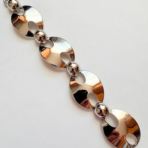 May include: A silver-toned bracelet featuring a series of large, oval links connected by smaller, spherical beads. The polished metal reflects light, creating a bright, shiny appearance. The bracelet has a clasp closure.
