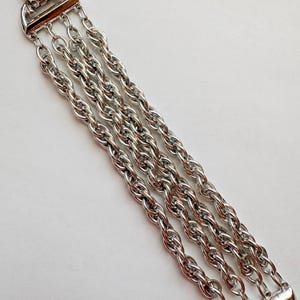 Puede incluir: Un brazalete plateado con cuatro cadenas paralelas y texturizadas. El brazalete tiene un cierre rectangular y una pieza superior decorativa. Las cadenas son de un material metálico brillante, sobre un fondo blanco. Mide unos 19 cm de largo.