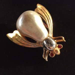 Pode incluir: Um broche dourado em forma de mosca com um corpo branco perolado grande e olhos de strass vermelhos.