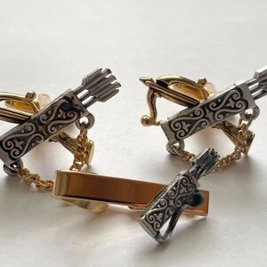 May include: A set of gold and silver cufflinks and tie bar featuring a bow and arrow design. The cufflinks are connected by a chain.