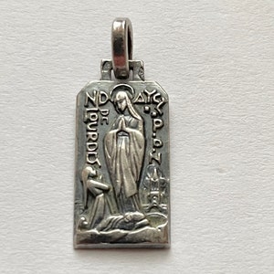 May include: Silver pendant featuring a depiction of the Virgin Mary, with the text "ND DE LOURDES" and "AYC" on the sides.
