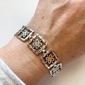 以下が含まれることがあります： 花柄のスクエアリンクが繰り返されるシルバーブレスレット。ブレスレットはシルバーとゴールドの2種類の金属で作られています。