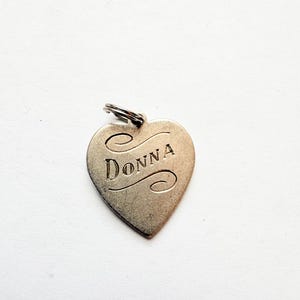 May include: A heart-shaped, silver-toned pendant with the name "Donna" engraved in a cursive font. The pendant has a small loop at the top for attaching to a chain or keyring. The background is a plain white surface.