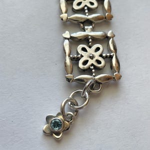 以下が含まれることがあります： 繊細なデザインのシルバーブレスレット。複数の正方形のリンクが連なり、各リンクには小さな花の模様が施されています。ブレスレットには、青い宝石が中心に付いた小さなチャームが付いています。