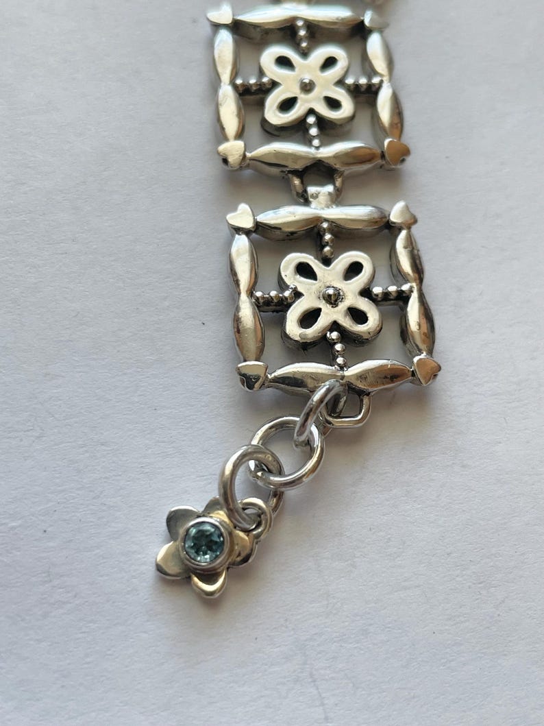 以下が含まれることがあります： 繊細なデザインのシルバーブレスレット。複数の正方形のリンクが連なり、各リンクには小さな花の模様が施されています。ブレスレットには、青い宝石が中心に付いた小さなチャームが付いています。