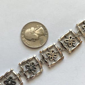 以下が含まれることがあります： 正方形と花が組み合わされた繊細なデザインのシルバーブレスレット。各正方形には、中央に石が埋め込まれた小さな花が飾られています。サイズ比較のために、アメリカ合衆国の25セント硬貨と一緒に写っています。