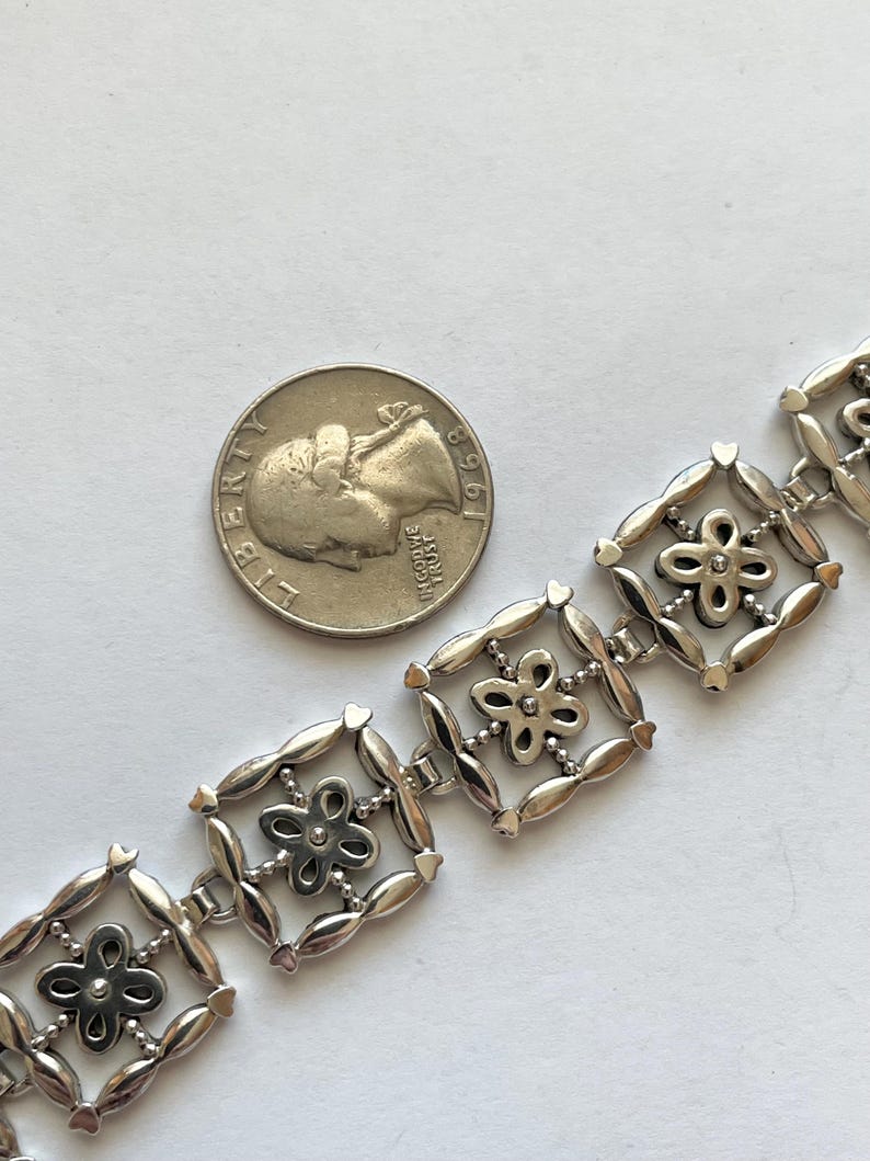 以下が含まれることがあります： 正方形と花が組み合わされた繊細なデザインのシルバーブレスレット。各正方形には、中央に石が埋め込まれた小さな花が飾られています。サイズ比較のために、アメリカ合衆国の25セント硬貨と一緒に写っています。