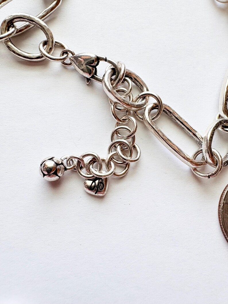 Puede incluir: Primer plano de una pulsera plateada con un dije en forma de coraz&oacute;n y un cierre de mosquet&oacute;n. La pulsera presenta una mezcla de dise&ntilde;os de eslabones ovalados y alargados, con un peque&ntilde;o dije de coraz&oacute;n y una bola decorativa.