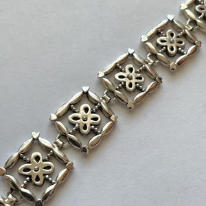 以下が含まれることがあります： 繊細な花のデザインが施されたシルバーのブレスレット。各リンクは、中央に小さな花がついた正方形のフレームになっています。ブレスレットは金属製で、磨かれた仕上げになっています。