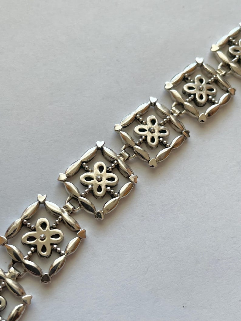 以下が含まれることがあります： 繊細な花のデザインが施されたシルバーのブレスレット。各リンクは、中央に小さな花がついた正方形のフレームになっています。ブレスレットは金属製で、磨かれた仕上げになっています。