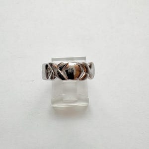 May include: A silver-colored ring with a woven design. The ring is displayed on a clear plastic stand against a white background. The ring's design features a series of interlocking shapes, creating a textured appearance. The ring is a piece of jewelry.