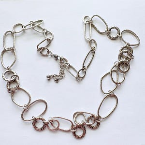 Puede incluir: Collar plateado con una variedad de formas de eslabones, incluyendo dise&ntilde;os ovalados y circulares. El collar tiene un acabado texturizado y un cierre de mosquet&oacute;n. La joya se muestra sobre una superficie blanca.