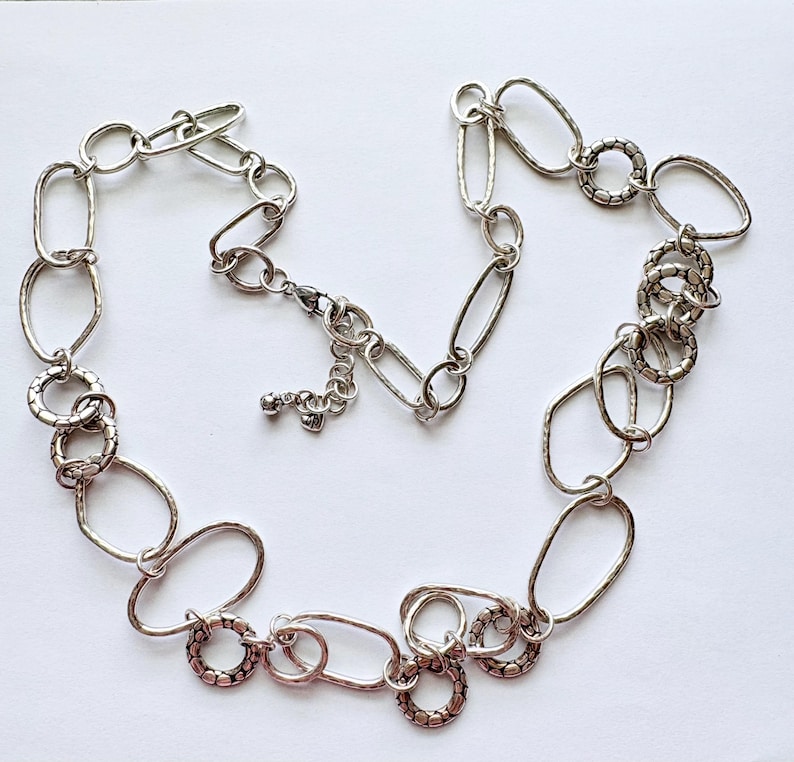 Puede incluir: Collar plateado con una variedad de formas de eslabones, incluyendo dise&ntilde;os ovalados y circulares. El collar tiene un acabado texturizado y un cierre de mosquet&oacute;n. La joya se muestra sobre una superficie blanca.