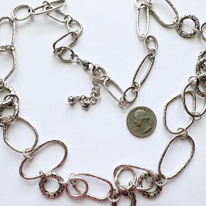 Puede incluir: Collar plateado con una mezcla de eslabones ovalados y circulares. Algunos eslabones tienen un aspecto texturizado y martillado. Se incluye una moneda para comparar el tama&ntilde;o, mostrando el dise&ntilde;o y los detalles.