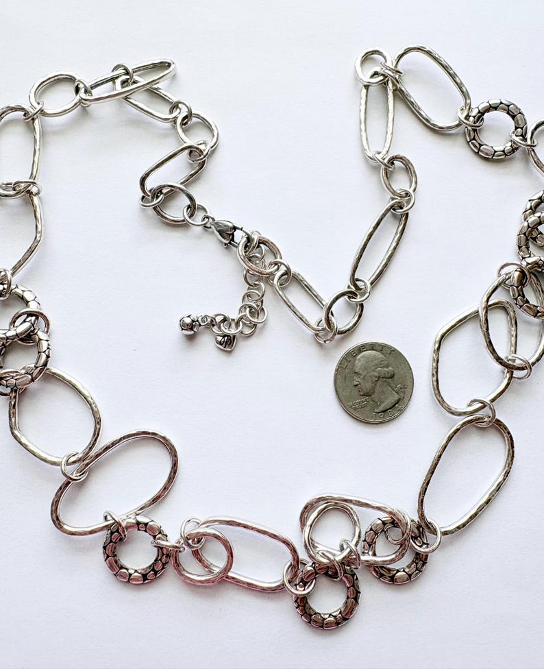 Puede incluir: Collar plateado con una mezcla de eslabones ovalados y circulares. Algunos eslabones tienen un aspecto texturizado y martillado. Se incluye una moneda para comparar el tama&ntilde;o, mostrando el dise&ntilde;o y los detalles.