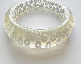 Puede incluir: Brazalete ancho y transparente con un tinte amarillo claro. El brazalete está decorado con adornos circulares de color plateado. El diseño es visible a través del material transparente, creando un efecto de capas. Un accesorio sencillo y elegante.
