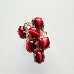 Könnte beinhalten: Ein silberner Ring mit einem Cluster aus sieben ovalen, tiefroten Edelsteinen. Die Edelsteine zeigen einen Katzenaugeneffekt. Die Fassung des Rings hat ein organisches, strukturiertes Design. Die Edelsteine sind etwa 1 cm lang.