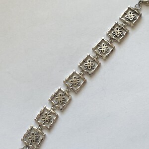 以下が含まれることがあります： 正方形のリンクが繰り返しパターンで並んだシルバーブレスレット。各リンクにはゴールドの花のデザインが施されています。