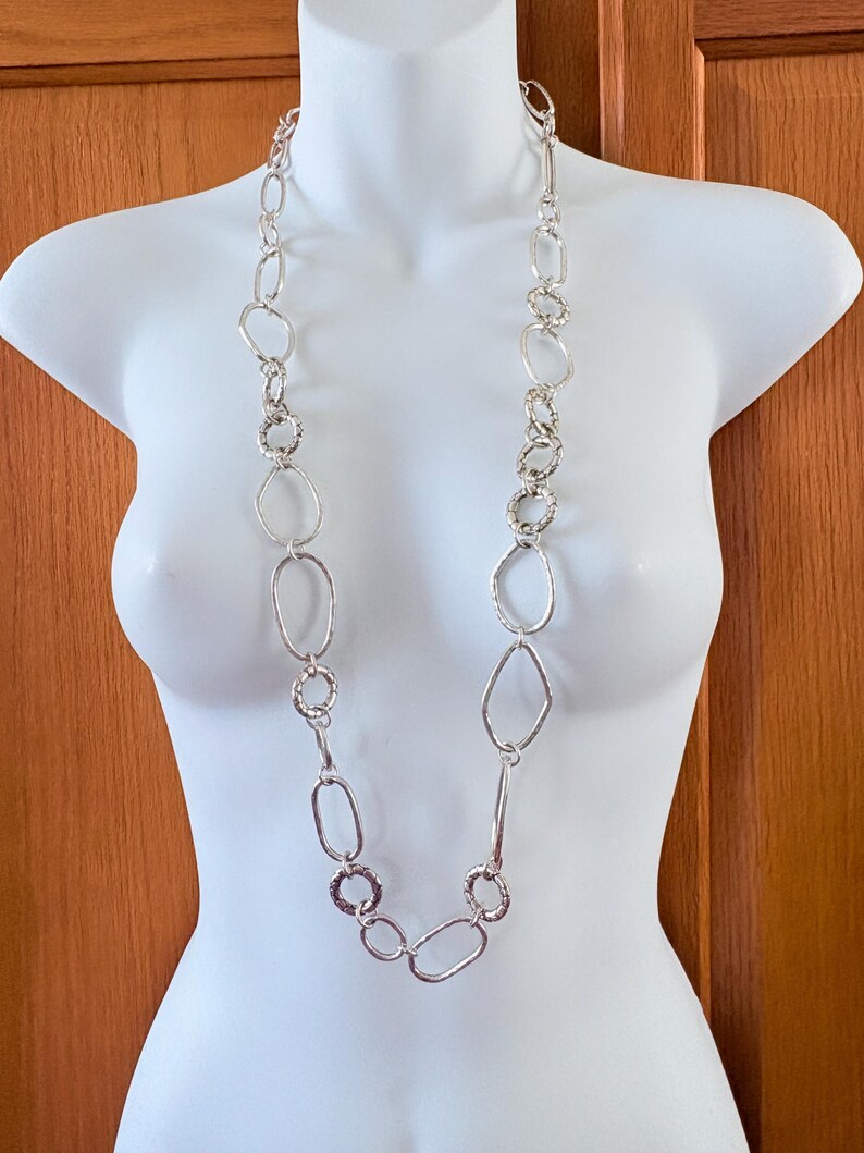 Puede incluir: Collar plateado con una variedad de formas geom&eacute;tricas, incluyendo &oacute;valos, c&iacute;rculos y rect&aacute;ngulos. El collar se exhibe en un maniqu&iacute; blanco, mostrando su longitud y dise&ntilde;o. Una pieza de joyer&iacute;a llamativa.