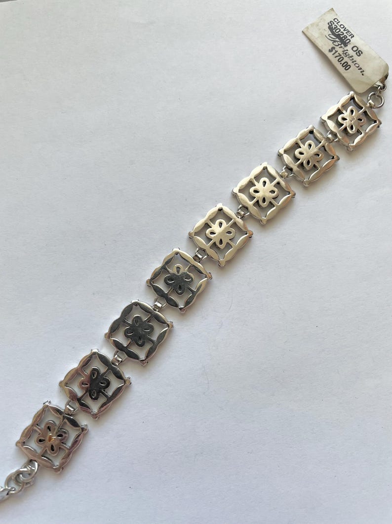 以下が含まれることがあります： 繊細で複雑なデザインのシルバーブレスレット。小さな正方形に花柄が繰り返されるパターンが特徴です。ブレスレットは、シルバーベースにゴールドのアクセントが施されたツートーンメタル製です。ブレスレットに付けられたタグには、「Clover OS 120.00 &euro;」と記載されています。