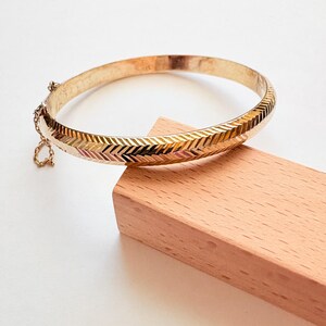 May include: A gold-colored bangle bracelet with a chevron pattern. The bracelet is resting on a small wooden block. The bracelet has a safety chain and clasp. The bangle is approximately 2.5 inches in diameter.