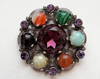 Könnte beinhalten: Eine silberne Brosche mit einem großen violetten Edelstein in der Mitte, umgeben von kleineren Edelsteinen in verschiedenen Farben, darunter Rot, Grün, Blau und Schwarz.