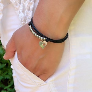 May include: A black leather bracelet with silver beads and a silver heart charm. The heart charm has a cut-out design.