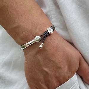 Peut inclure: Un bracelet jonc en argent avec un cordon en cuir marron attaché. Le cordon est orné d'un charme en forme de fleur argentée et de deux petites perles argentées.