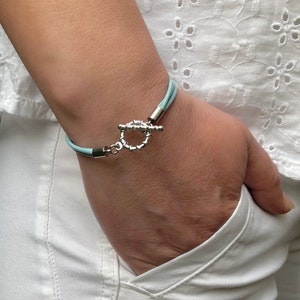 May include: A light blue leather bracelet with a silver toggle clasp. The bracelet is worn on a person's wrist.
