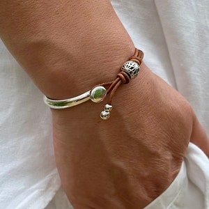 May include: A brown leather bracelet with a silver charm and two silver beads. The bracelet is tied with a knot and has a silver clasp.