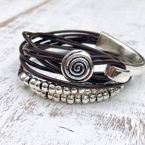 Könnte beinhalten: Ein braunes Lederarmband mit einem silbernen Spiral-Anhänger und silbernen Perlen.
