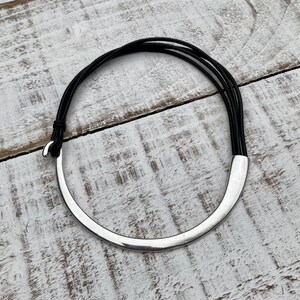 K&ouml;nnte beinhalten: Eine silberne Choker-Kette mit einem geschwungenen Design und einem schwarzen Lederband.