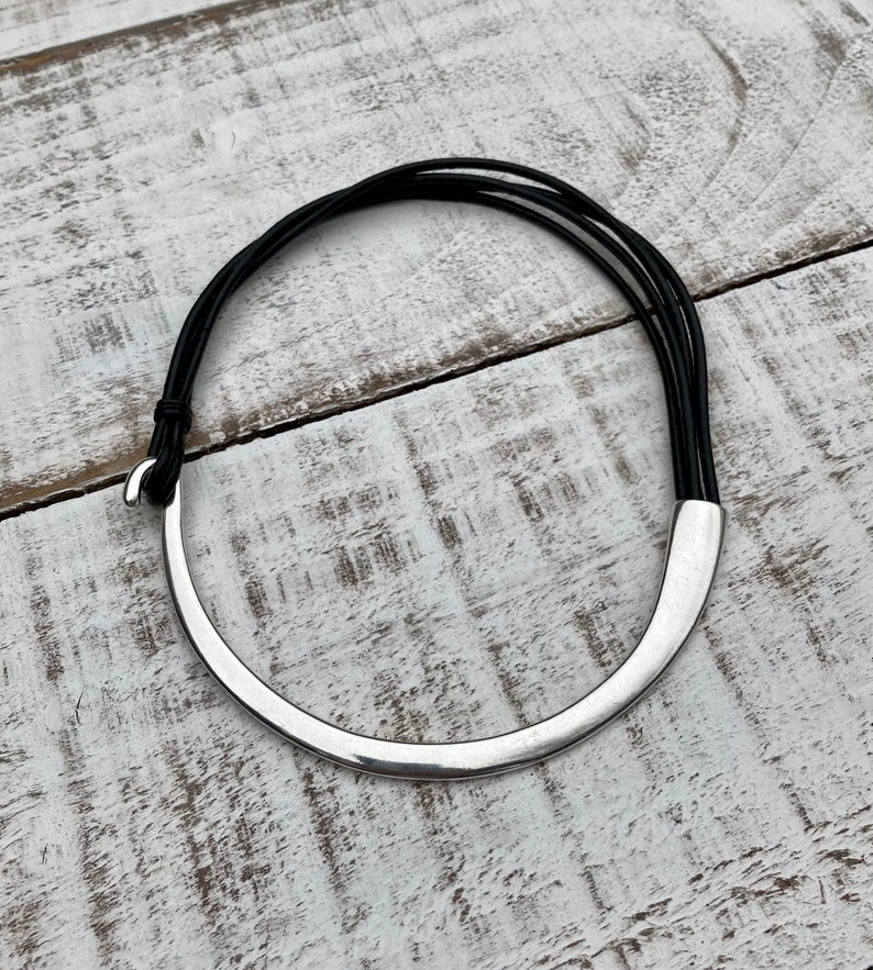 K&ouml;nnte beinhalten: Eine silberne Choker-Kette mit einem geschwungenen Design und einem schwarzen Lederband.