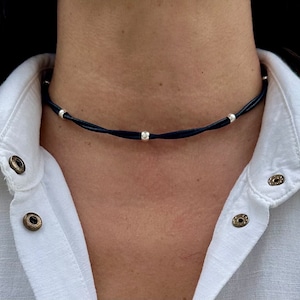 May include: A black leather choker necklace with silver beads. The necklace is worn by a person with a white shirt.
