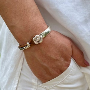 Peut inclure: Un bracelet en cuir marron avec des perles argentées et un charme en forme de fleur argentée. Le bracelet est porté au poignet.