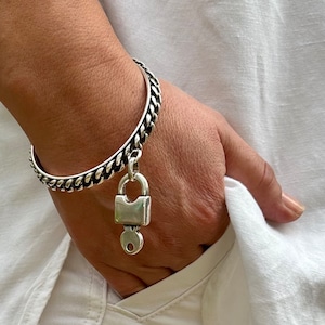 May include: A silver chain bracelet with a padlock charm and a key. The bracelet is on a person's wrist.
