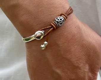 May include: A brown leather bracelet with silver beads and a silver charm. The bracelet is tied with a knot and has a small silver bead on the end.