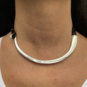 K&ouml;nnte beinhalten: Eine silberne Choker-Kette mit einem geschwungenen Design und schwarzen Lederriemen.