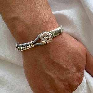 May include: A silver bangle bracelet with a grey leather strap and a silver flower charm. The bracelet is on a person's wrist.