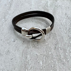 May include: A brown leather bracelet with a silver hook clasp. The bracelet is double-layered and has a silver accent on the strap.