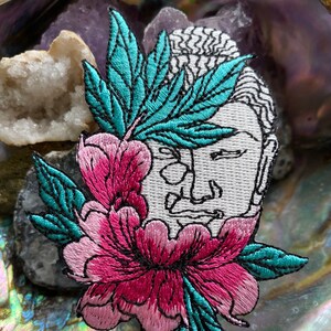 May include: Embroidered patch featuring a white Buddha head with black outlines, adorned with a pink and teal flower and teal leaves. The patch is set against a backdrop of crystals and a pearlescent surface.