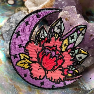 May include: Embroidered patch featuring a purple crescent moon with small stars and a central floral design. The flower has red, pink, and yellow petals, with black outlining. The patch is likely for clothing or accessories.