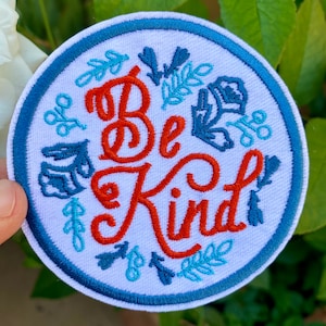 May include: Round patch with the words "Be Kind" in orange script, surrounded by blue floral designs on a white background. The patch has a blue border and is likely an iron-on or sew-on patch.