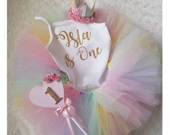 May include: A rainbow tulle tutu with a white onesie that says "Isla is One" in gold glitter. A pink heart-shaped wand with a gold glitter number one and a pink bow. A white crown with a gold glitter number one and pink flowers.