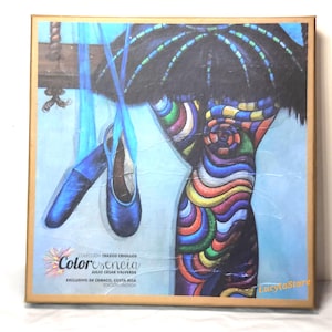 May include: A square, framed artwork featuring a ballet theme. It depicts a colorful, stylized figure with a dark umbrella-like shape above. Two blue ballet shoes hang from a wooden beam. The artwork includes the text "Color esencia" and "Julio Cesar Valverde."